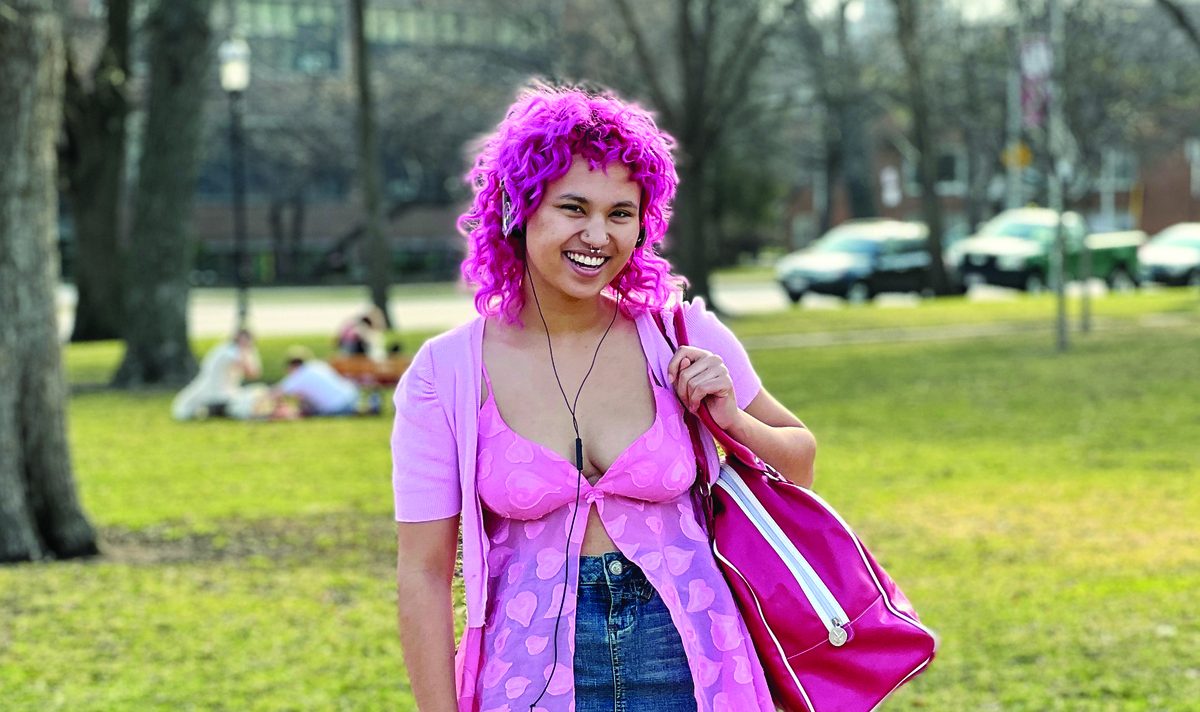a person with magenta colored hair wearing a pink top and blue denim skirt with hot pink tote bag and shoes; they are standing on a sidewalk near a stretch of green grass
