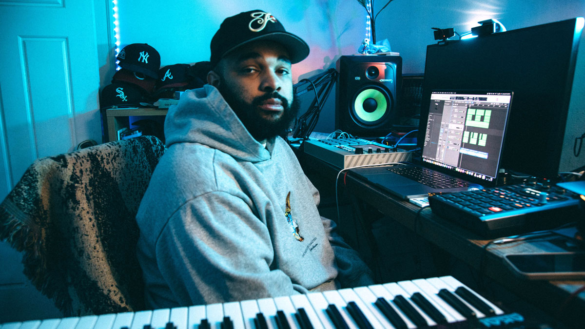Thelonious Martin sits in his blue-lit home studio in a hoody and brimmed cap, surrounded by keyboards, computers, samplers, drum machines, and monitor speakers.