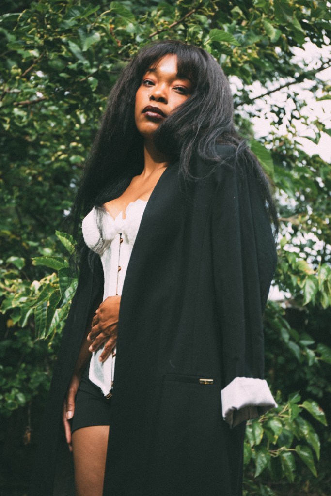 Loona Dae looks down at the camera against a backdrop of green leaves, with a long black coat draped over her shoulders and a white corset-style top