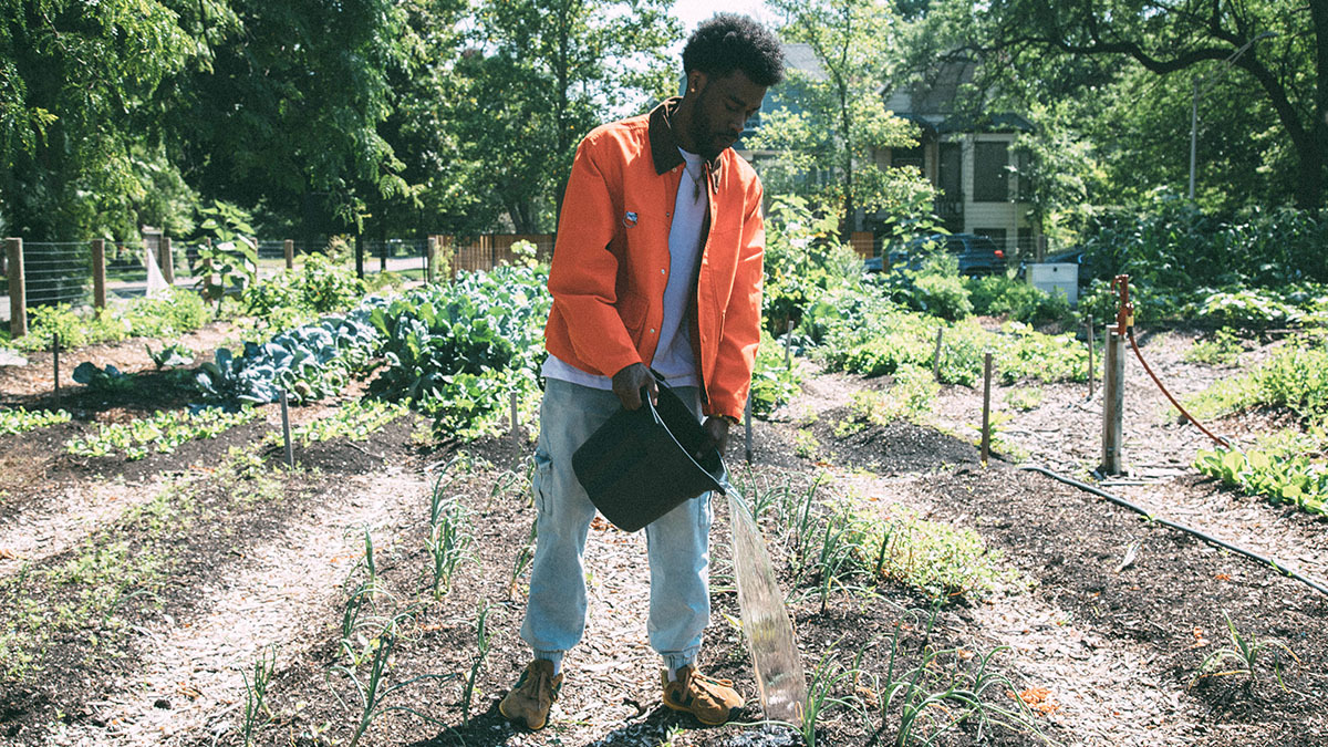 a man in jeans and an orange jacket stands in a garden plot with a large black plastic bucket