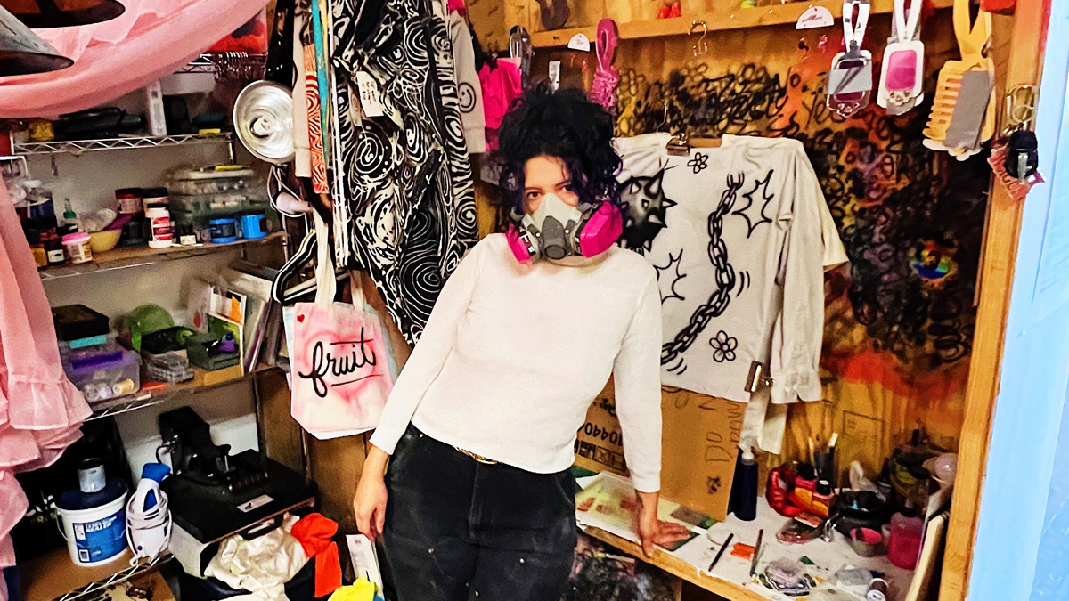 Izzi Vasquez leans against a work desk in a cluttered and colorful art studio, wearing bright red boots and a magenta-and-white respirator mask