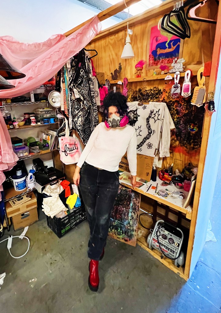 Izzi Vasquez leans against a work desk in a cluttered and colorful art studio, wearing bright red boots and a magenta-and-white respirator mask