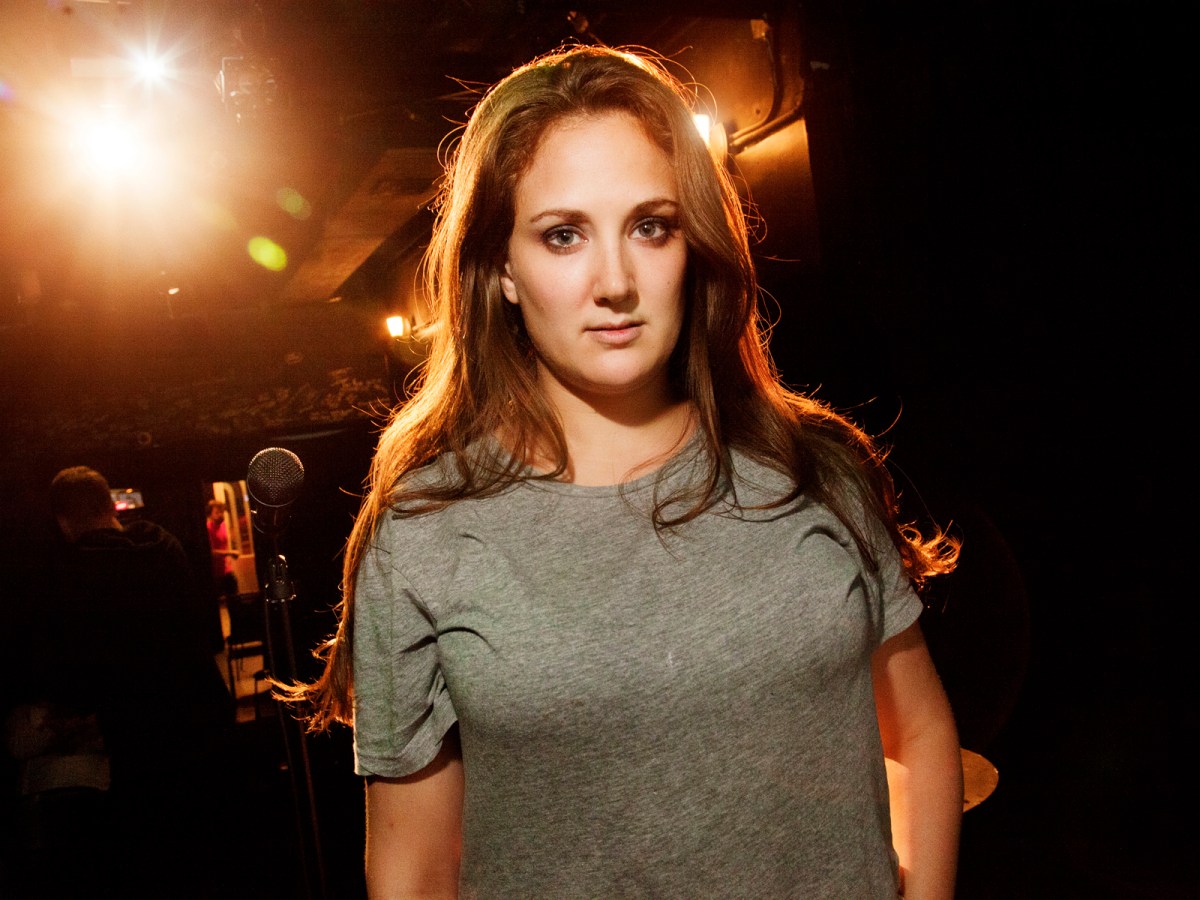Jacqueline Novak (long brown hair, brown eyes) wearing a gray T-shirt and backlit onstage standing in a