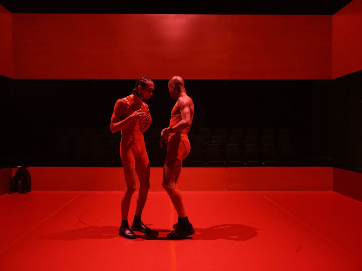 Two Black men stand facing each other. The man on the left has short braids. The man on the right is bald. The stage is mostly bare except for a stool in the far upper left corner. The setting is reminiscent of a boxing ring, and the playing area is drenched in red light.