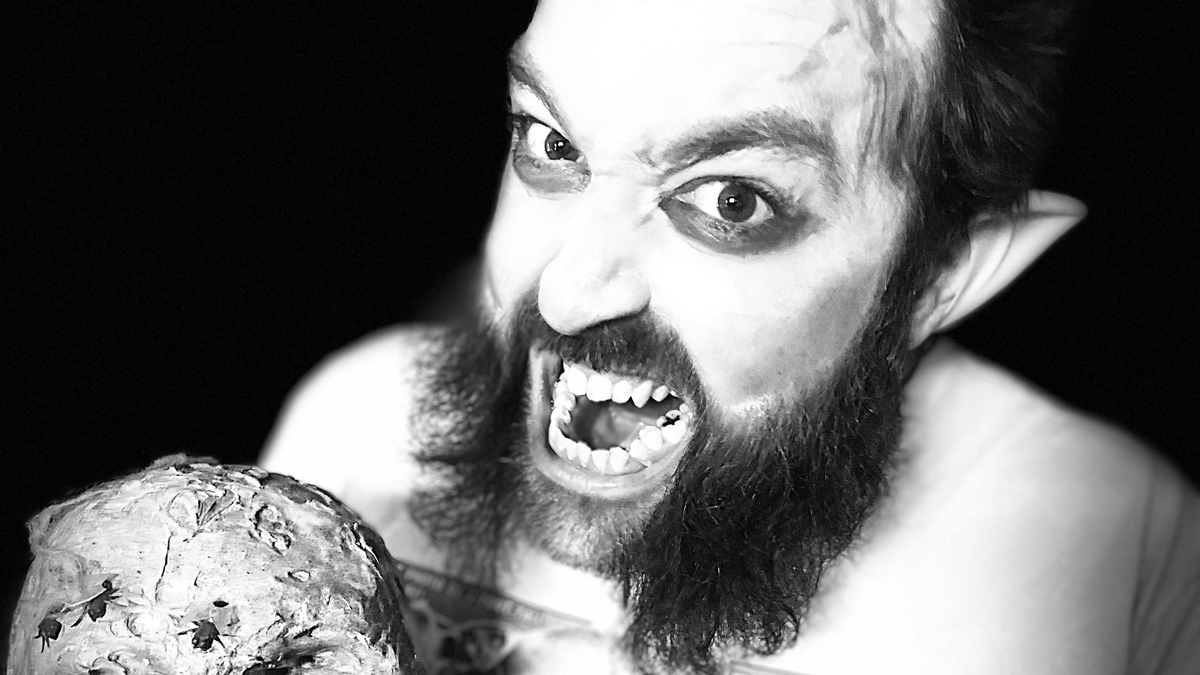 a black-and-white portrait of Josh Pietrowski snarling at the camera with vampire makeup, pointed prosthetic ears, and a widow's peak; he holds a decorated skull sculpture and wears a T-shirt of the famous Weekly World News issue that introduced the Bat Boy character