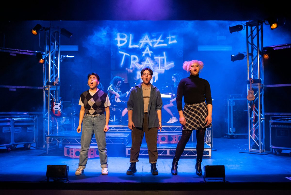 Three actors from BoHo Theatre's production of tick, tick . . . BOOM! stand onstage in front of a projection reading "Blaze a Trail."