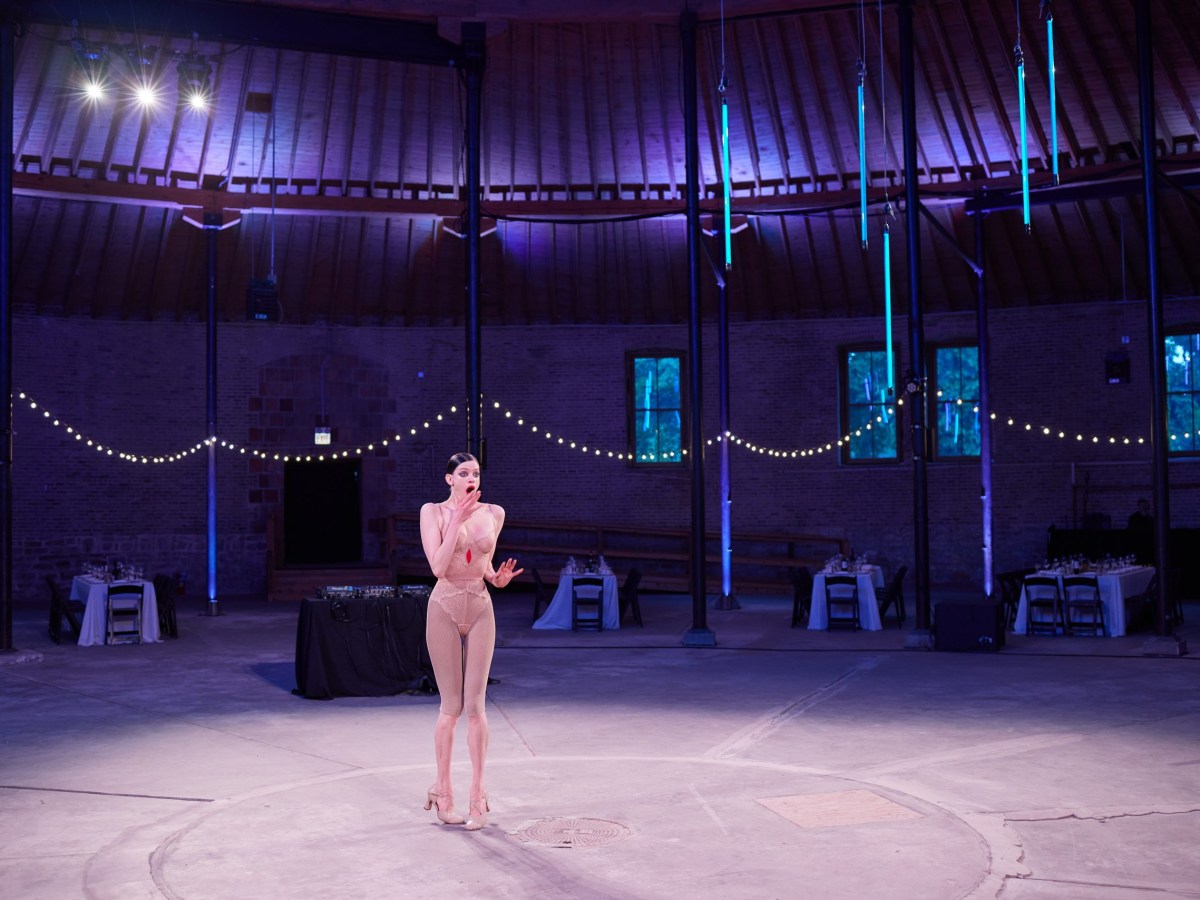 Lily is dressed in layers of nude lingerie. She stands in the center of the Rotunda, which is empty save for tables behind her. She is lit by a spotlight.