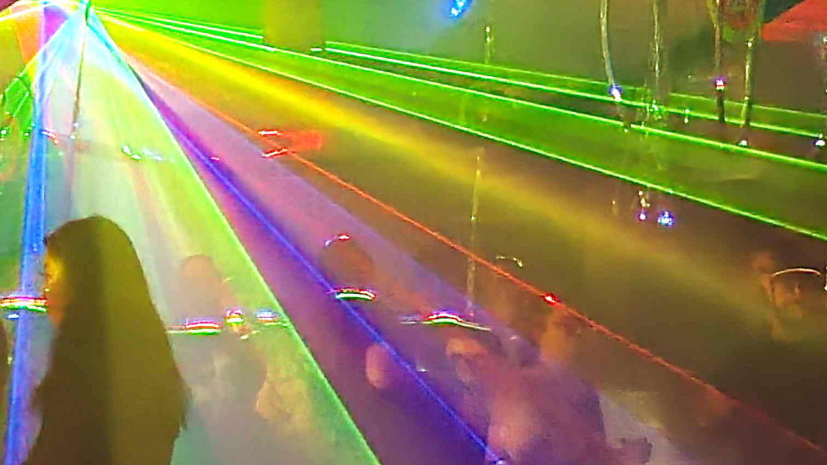 rays of colored light radiate across a darkened room filled with people at a small dance concert