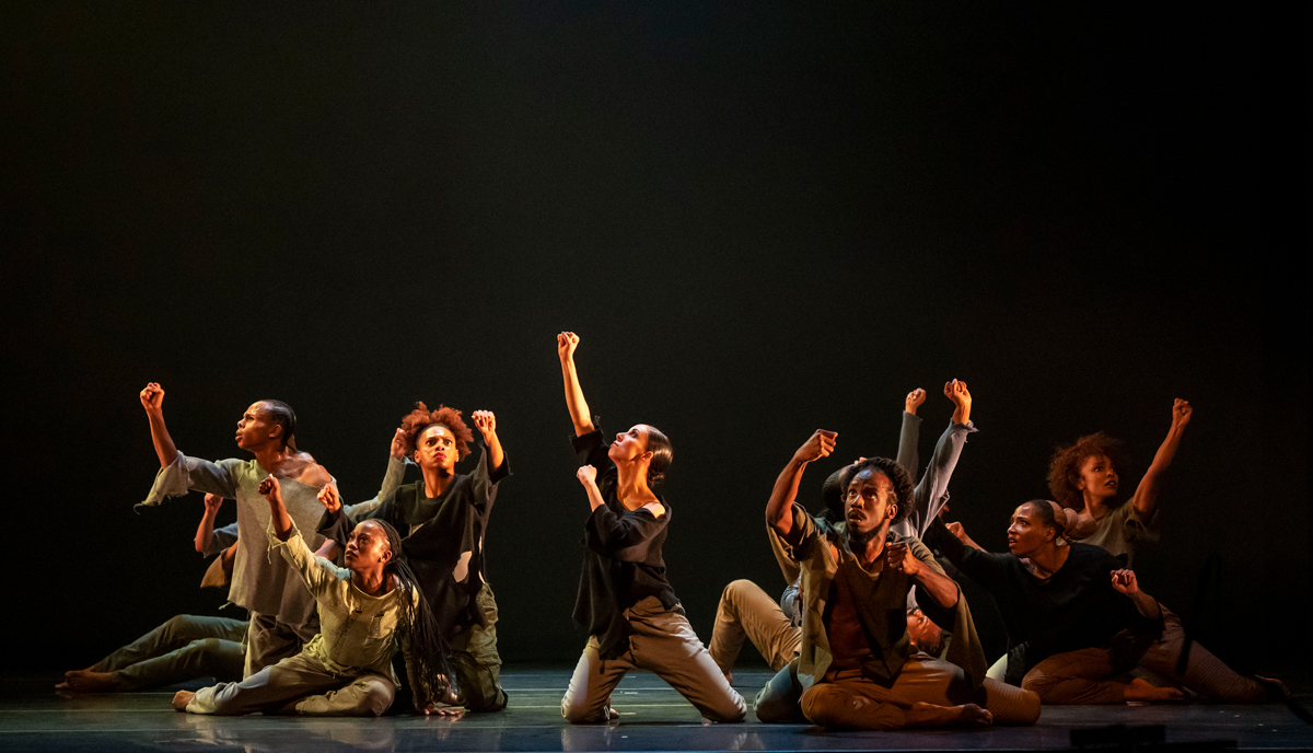 A group of over a half dozen dancers, kneeling and sitting onstage with their fists upraised.