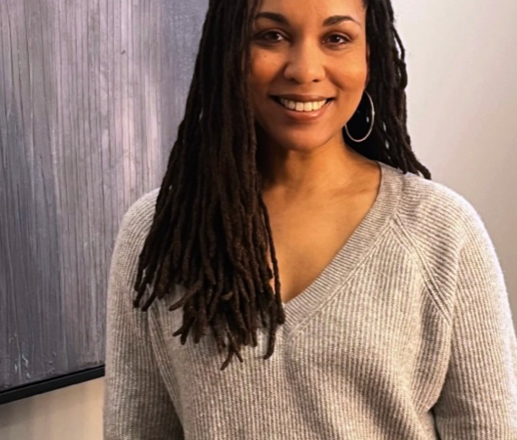 Meredith Sutton, a Black woman with long braided hair, is photographed from the waist up. She is wearing a gray V-neck sweater and silver hoop earrings. In the background on the left there appears to be some sort of screen or blackboard.