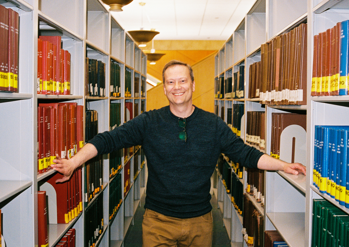 a white man poses for a photo between two library bookshelves