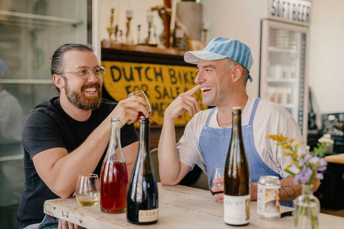 two laughing men sit at a wooden table with wine bottles and glasses