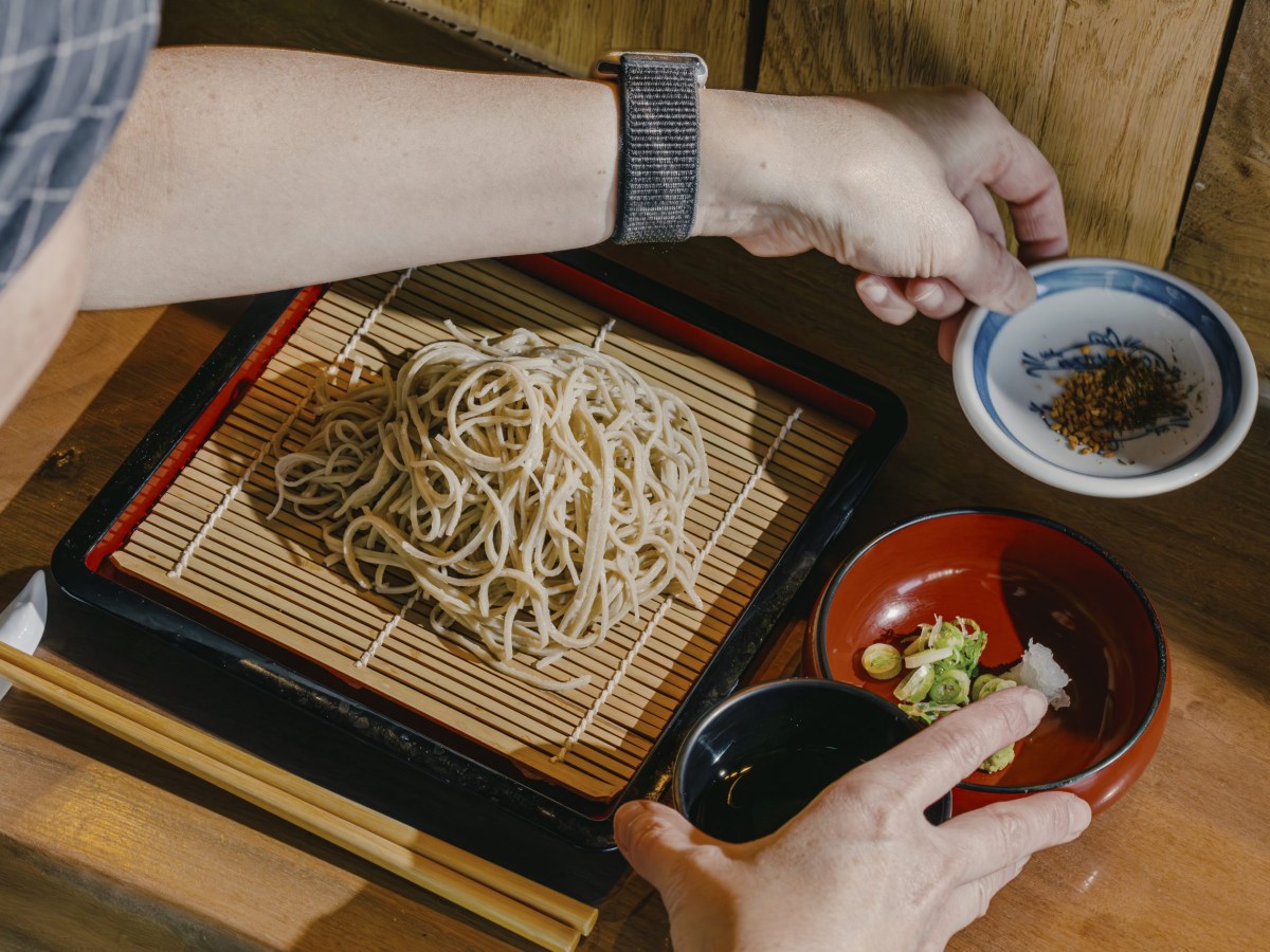hands arrange soba noodles on a square mat next to chopsticks and three small dishes with sauces, spices, and green onions