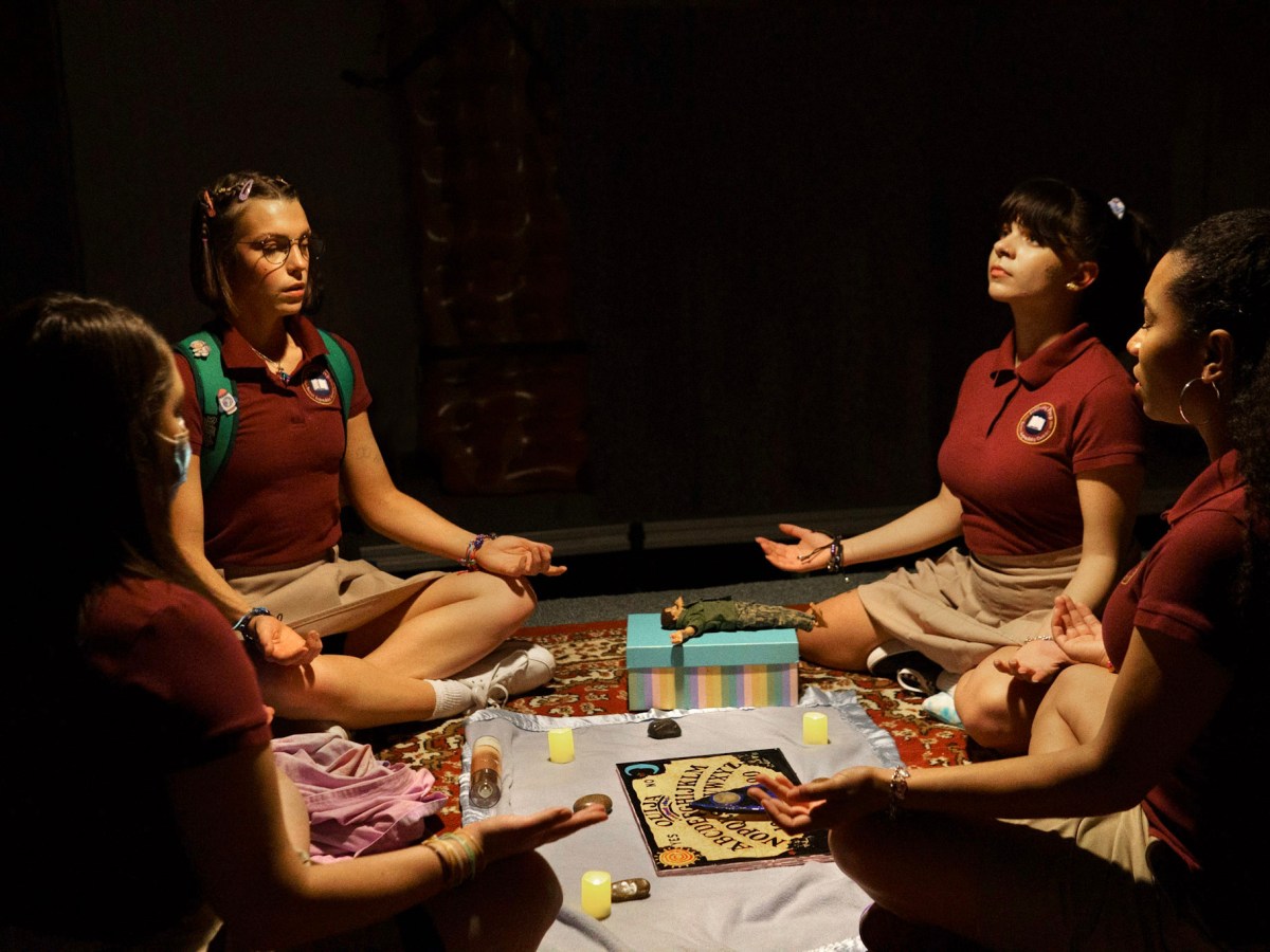 Four girls of various ethnicity and race, dressed in school uniforms, sit cross-legged around a Ouija board.