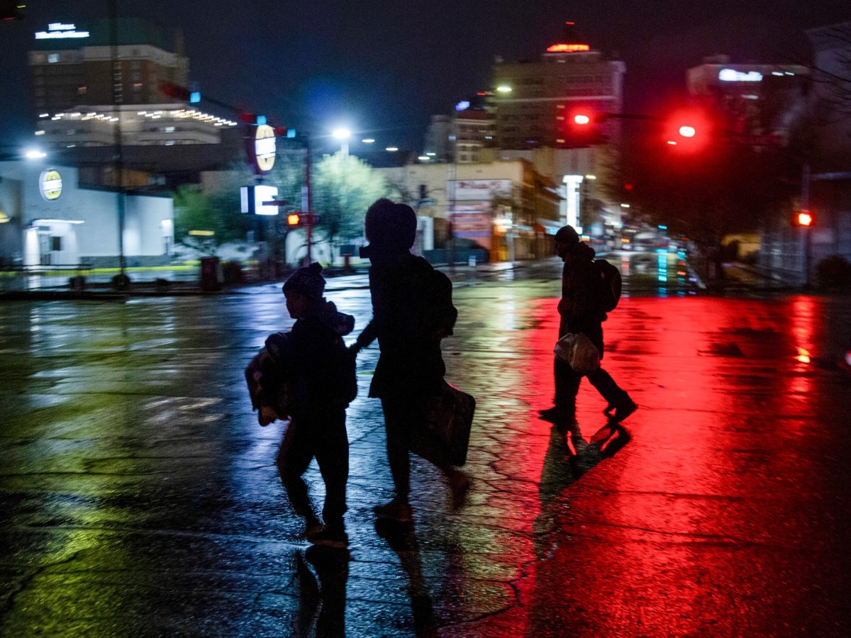 A family crosses a wet city street at night. The street is light red from a stoplight and blue and green from the neon lights of businesses.
