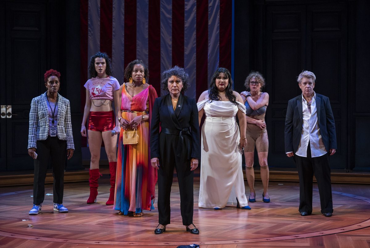Seven women of different ages, races, and ethnicity, all in some state of dishevelment, stand in a line onstage. Part of a large American flag is visible hanging behind them.