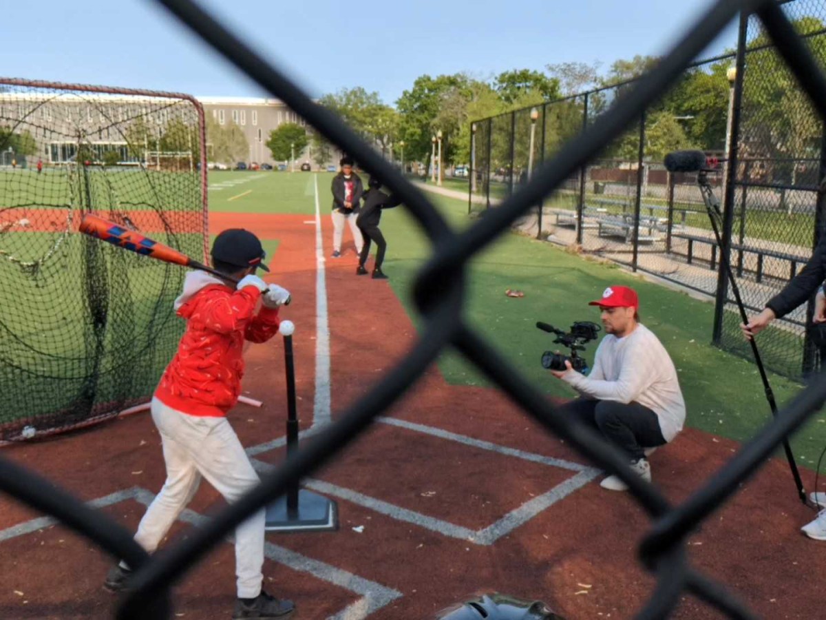 through a chainlink fence, a shot of a cameraman recording a young baseball player at bat