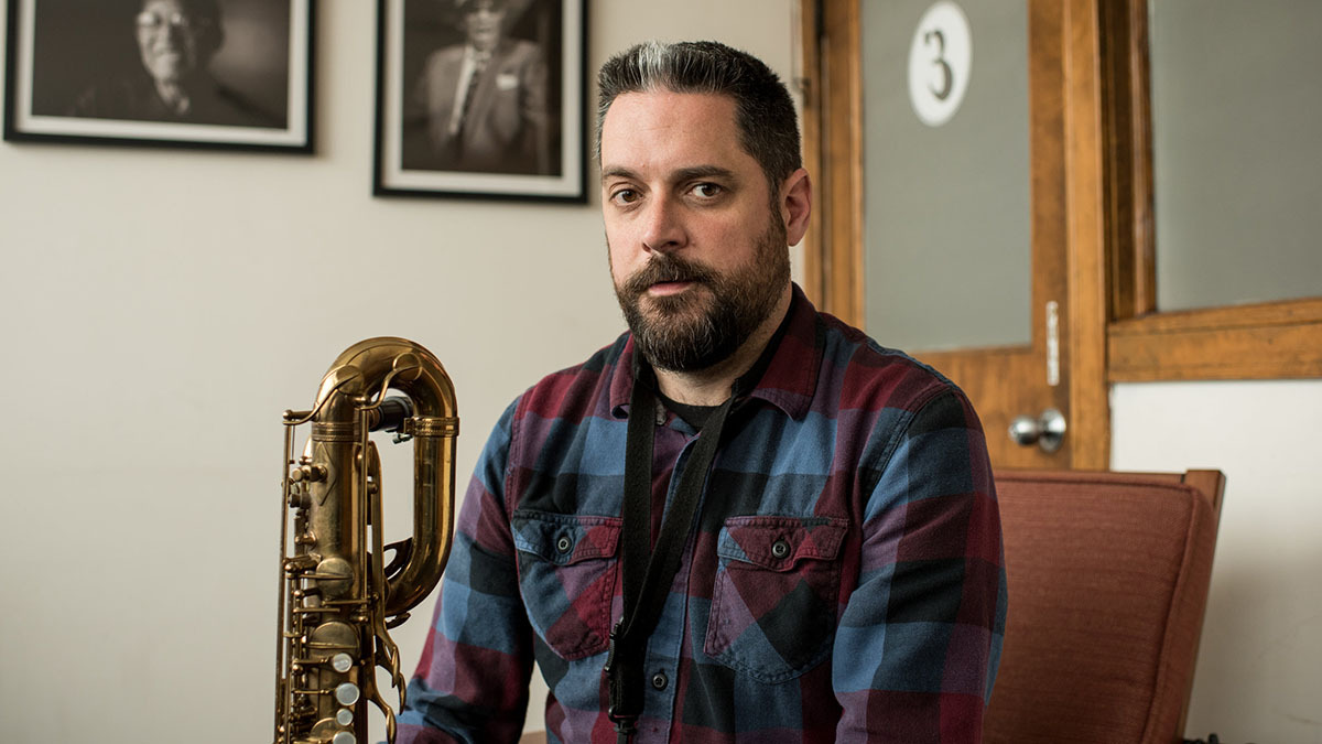 Dave Rempis looks into the camera while seated in an office, the neck of a baritone saxophone visible in the frame to his left