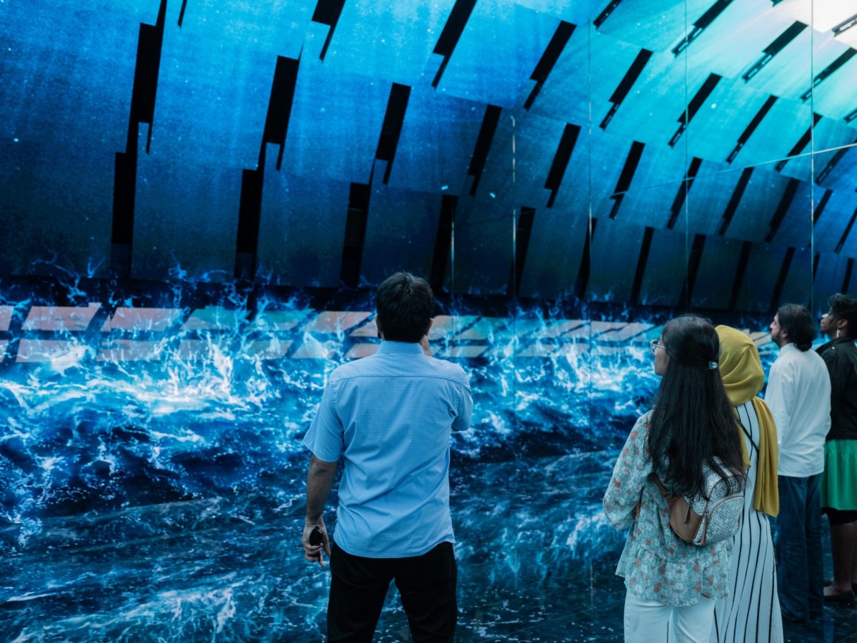 A small crowd of people stand in front of a wall playing a video of an ocean wave crashing.