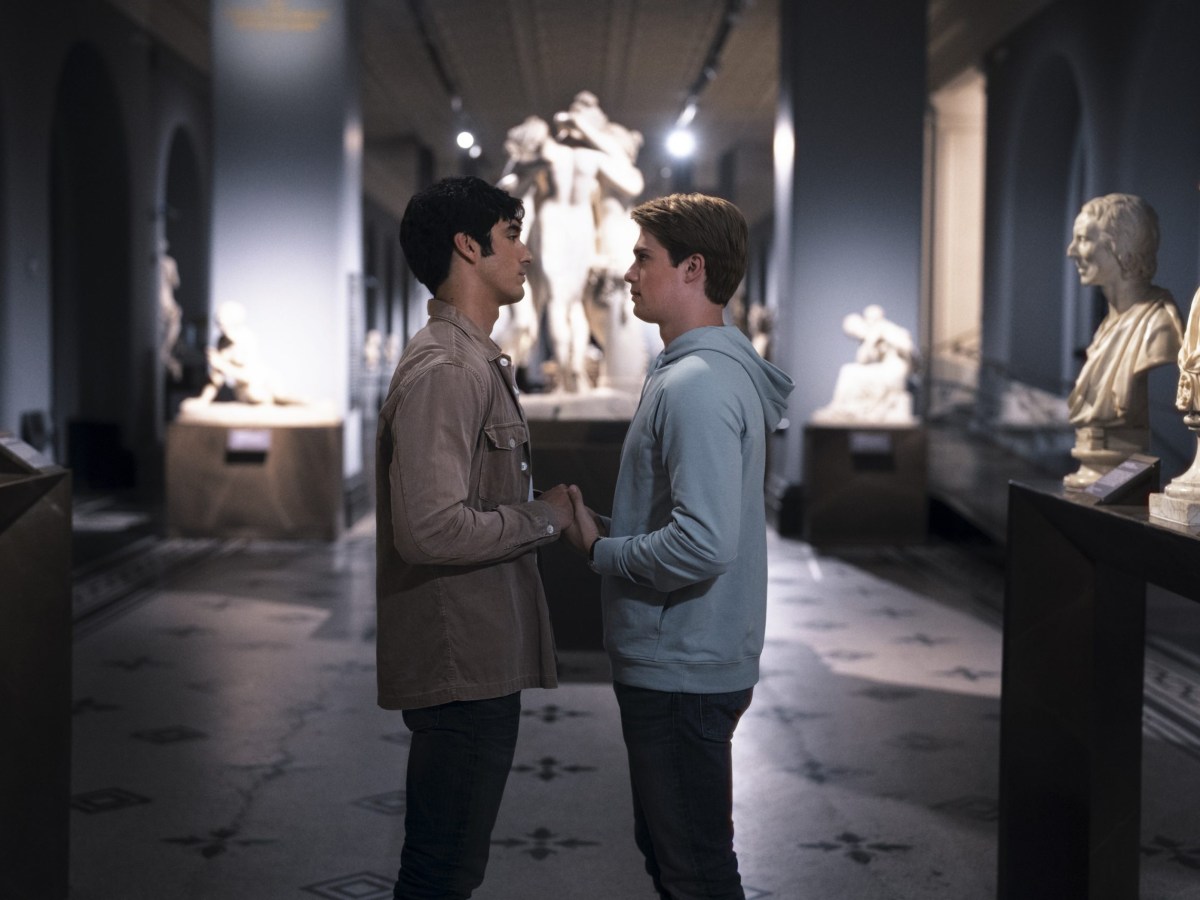 two young men hold hands in a museum full of sculptures and statues
