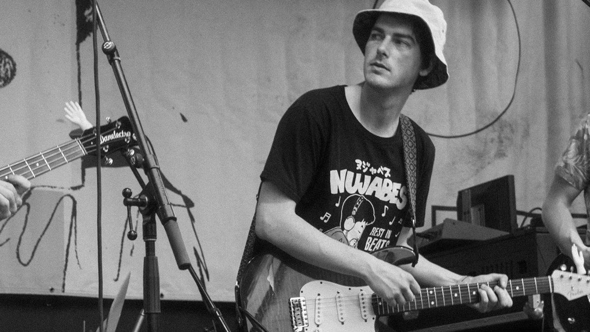 a black-and-white photo of Ryan Deffet playing guitar onstage, wearing a light-colored bucket hat and a dark Nujabes T-shirt