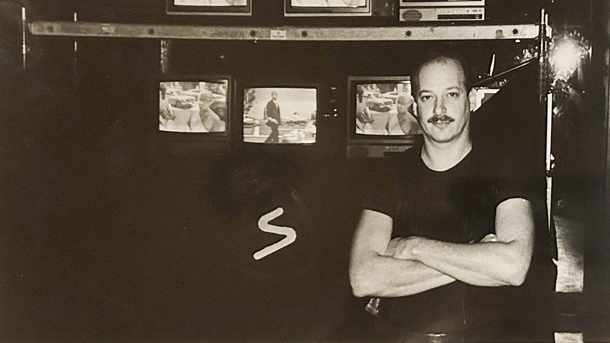 a black-and-white photo of Shelley Howard, facing the camera with his arms folded, with an array of seven turned-on TV sets behind and above him, showing two different images
