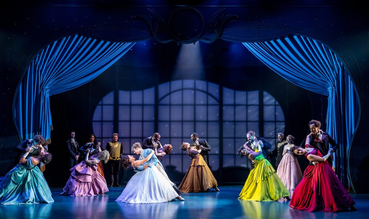 Women in ball gowns and men in fancy suits dance in a line across the stage.