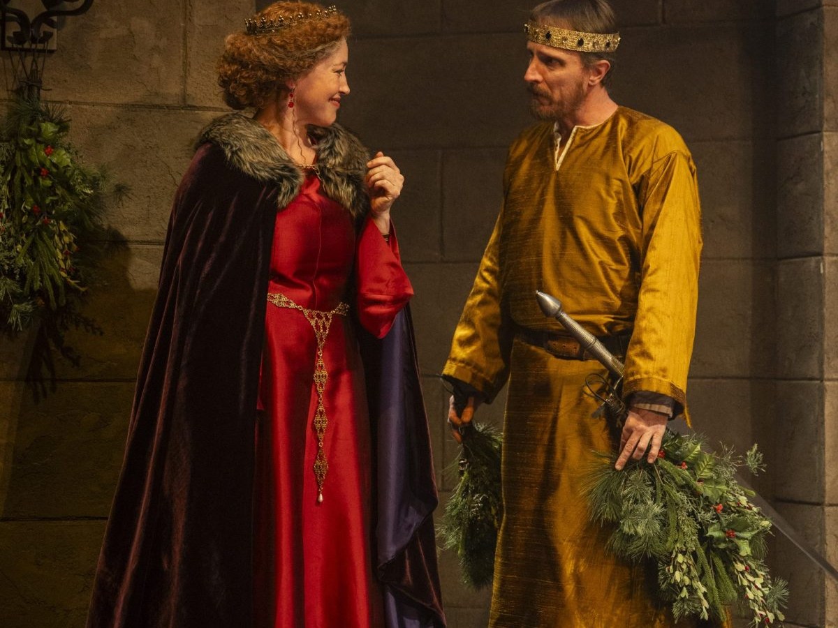 A woman in a long red dress and fur-trimmed cloak stands left. On the right is a man in a gold gown and a crown, carrying an evergreen branch.