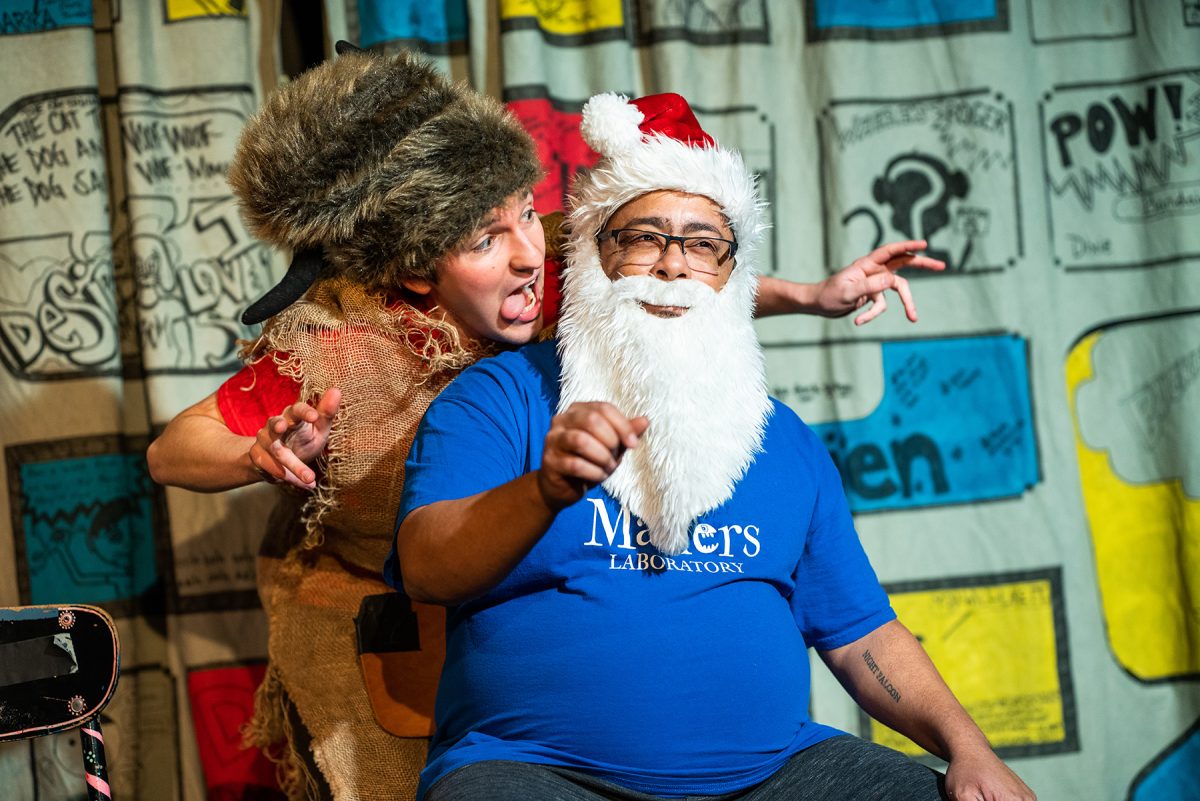 Linsey Falls, wearing a blue PlayMakers T-shirt and a Santa hat and beard, sits center. A man in a coonskin cap and burlap smock stands behind him, leaning in. Behind them is a backdrop with comic strips.