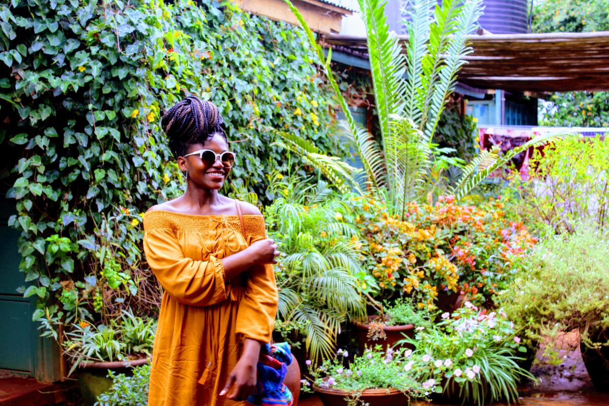 a Black woman wearing an orange dress and sunglasses stands in the middle of a lush green garden