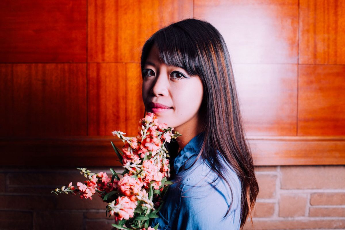 poet and writer Jane Wong posing in front of a wooden wall, holding a bouquet of flowers