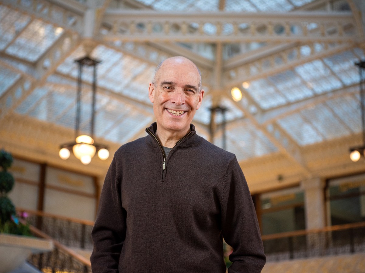 a bald white man in a quarter-zip sweater poses for a phot, with an impressive building interior behind him
